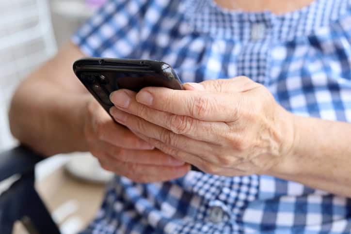 Elderly woman in checkered dress sitting with smartphone in chair, mobile phone in wrinkled female hands closeup