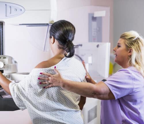 A mature African-American woman in her 40s wearing a hospital gown, getting her annual mammogram. She is being helped by a technologist, a blond woman wearing scrubs.