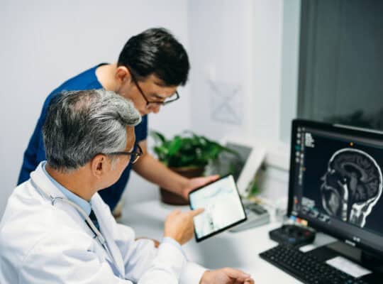 Two Asian male doctors are using digital tablet computer to discuss,  and monitors showing brain scans results In control room.