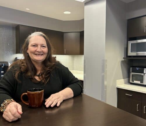 Woman sitting at modern kitchen counter holding coffee mug, smiling at camera