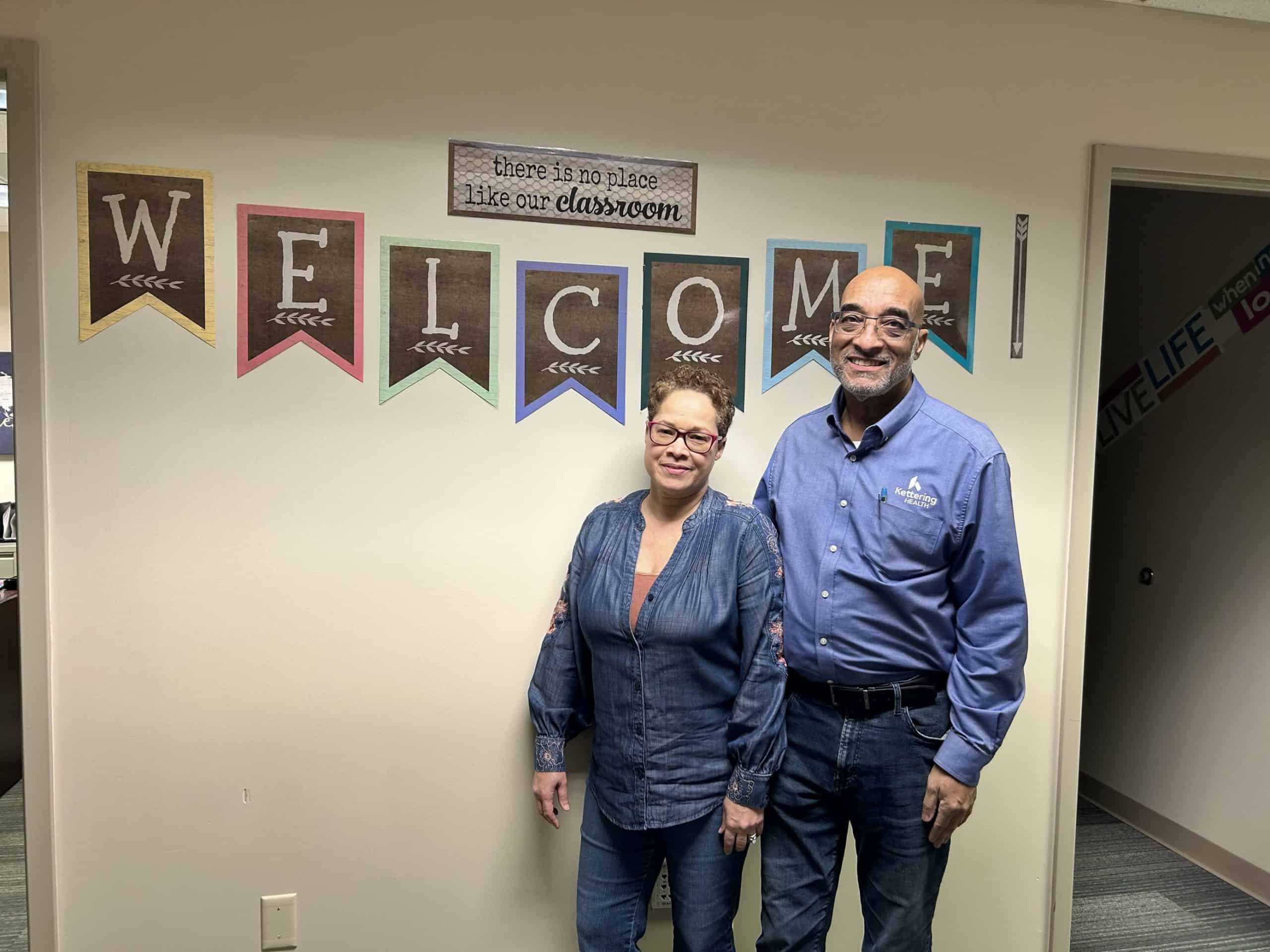 Two healthcare professionals standing together in classroom with welcome banner overhead
