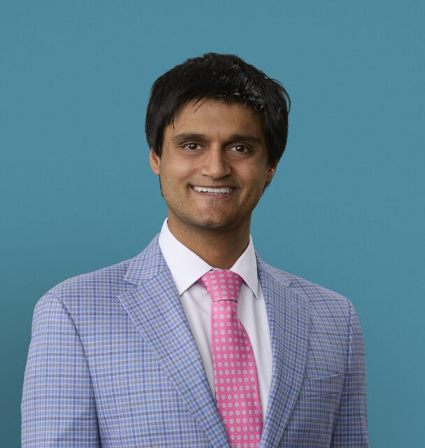 Professional headshot of Dr. Amrit Thakur in light blue checkered suit with pink tie