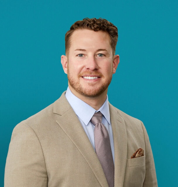 Professional headshot of Dr. Cord Gothard in tan suit against teal background