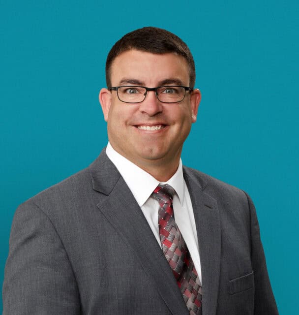 Professional headshot of Dale Guinther, APRN-CNP in gray suit with glasses smiling