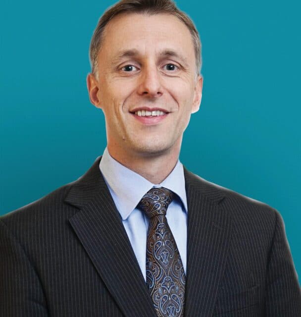 Professional headshot of Dr. Gregory F. Egnaczyk in dark pinstripe suit against teal background
