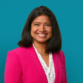 Professional headshot of Dr. Heena Santry in bright pink blazer against teal background