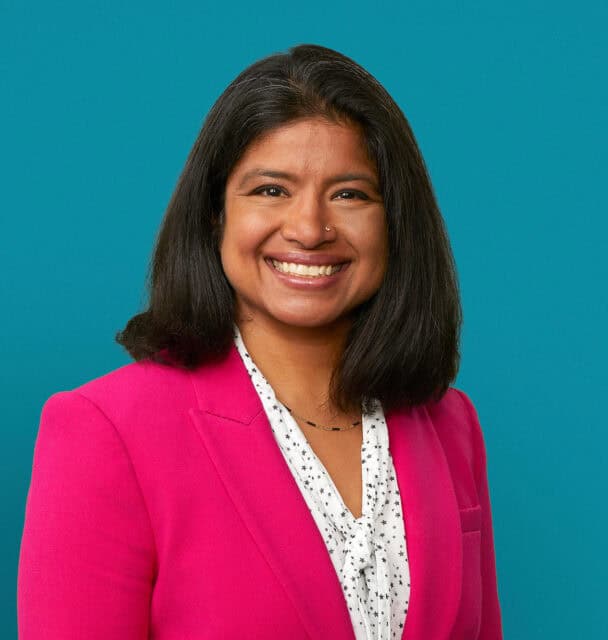 Professional headshot of Dr. Heena Santry in bright pink blazer against teal background