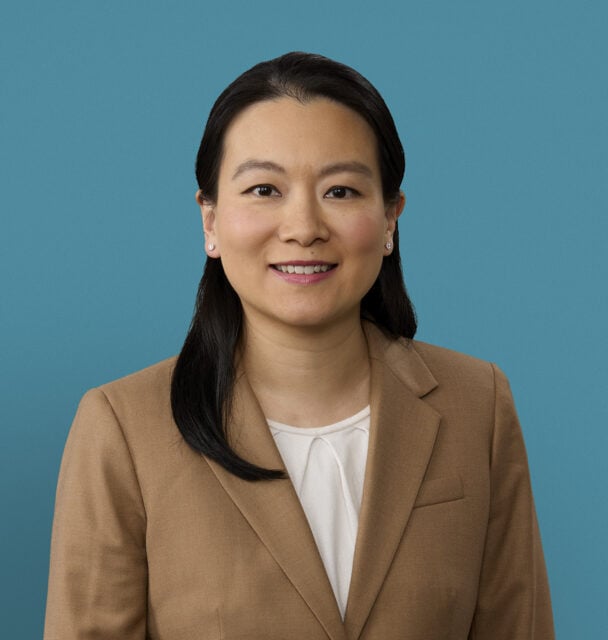 Professional headshot of Dr. Jing Li Huang in tan blazer against blue background