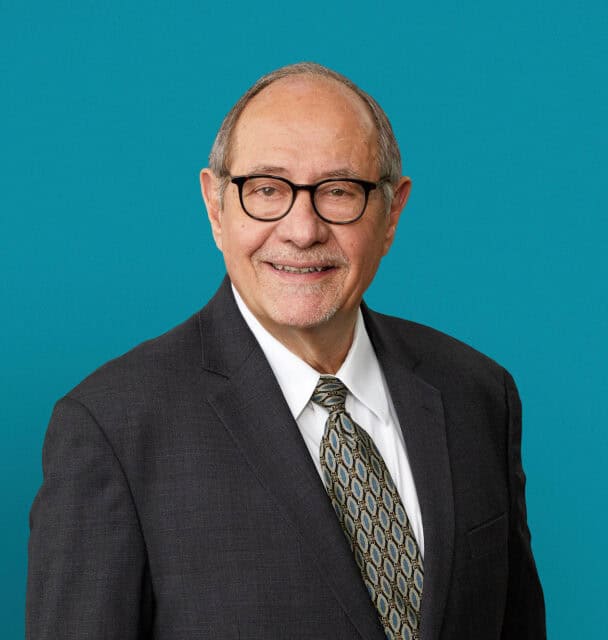 Professional headshot of John Hollon, MD wearing glasses and suit against teal background