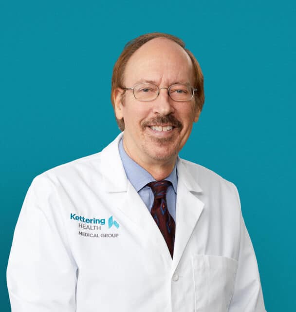 Professional headshot of John R. Bullmaster, MD in white coat against teal background