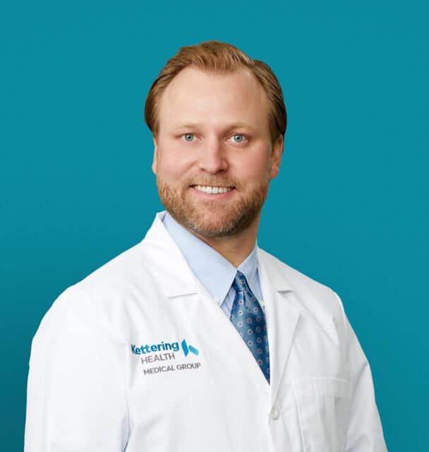 Professional headshot of Dr. Joshua A. Ahier in white coat against teal background