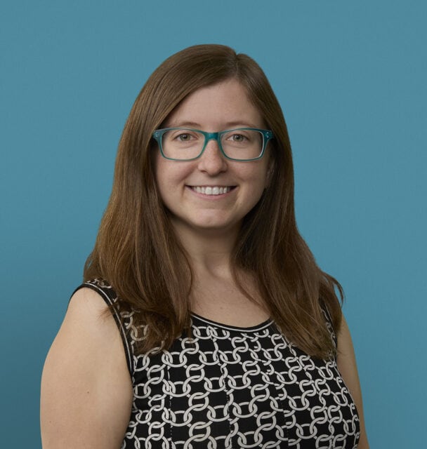 Professional headshot of Kate Meizlish, MD smiling in black patterned top with teal glasses