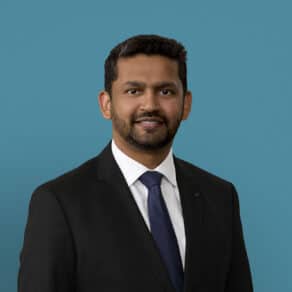 Professional headshot of Dr. Nazir Ahmed in dark suit with blue tie against teal background