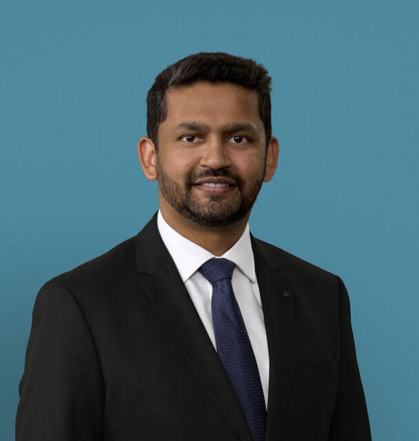 Professional headshot of Dr. Nazir Ahmed in dark suit with blue tie against teal background