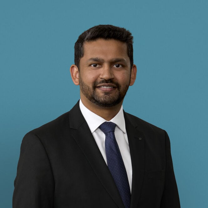 Professional headshot of Dr. Nazir Ahmed in dark suit with blue tie against teal background
