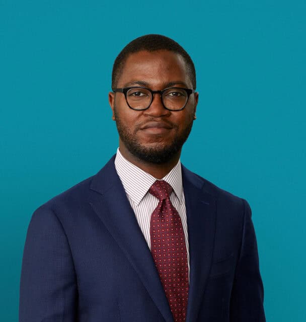 Professional headshot of Dr. Olaoye Jegede in navy suit against teal background