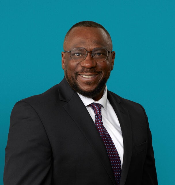 Professional headshot of Dr. Rory Lewis in dark suit and tie against teal background