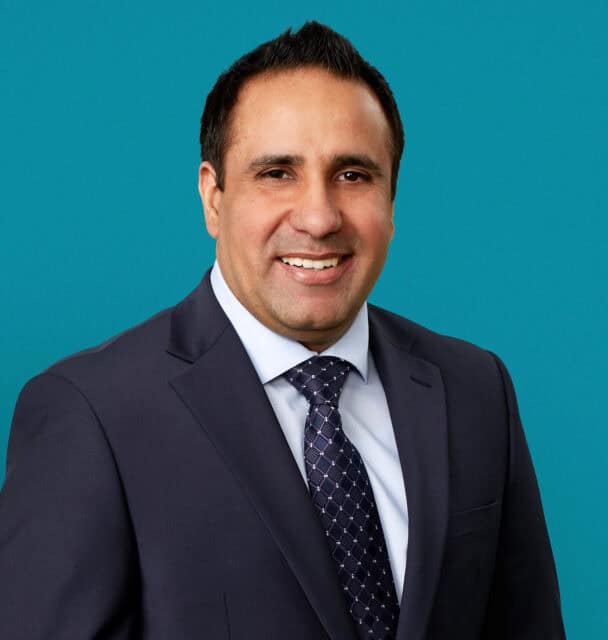 Professional headshot of Dr. Sukhchain Singh in dark suit against teal background