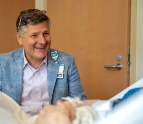 Man in blue blazer smiling while sitting beside hospital bed with patient