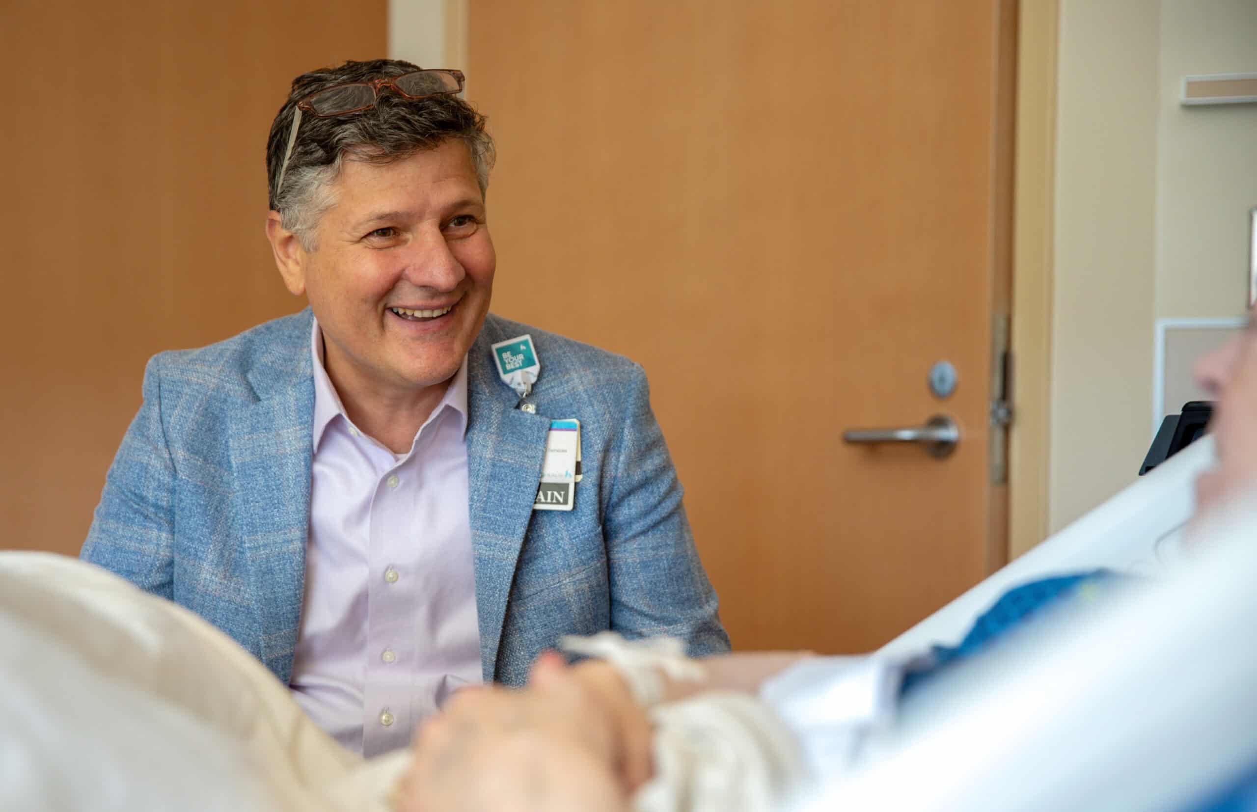 Man in blue blazer smiling while sitting beside hospital bed with patient