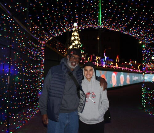 Two people posing together under colorful Christmas lights and holiday decorations at night